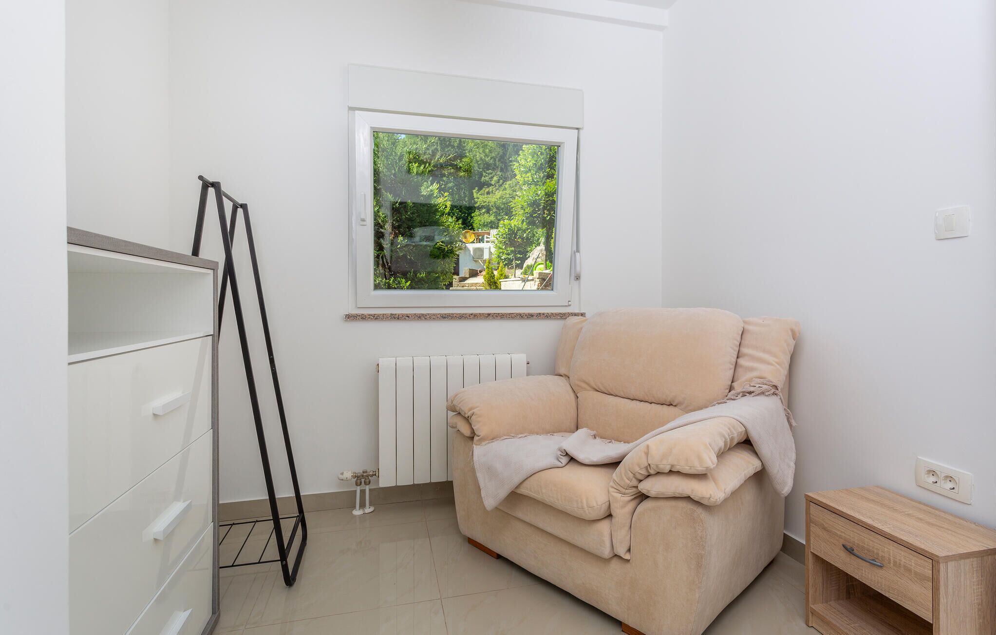 Habitación minimalista con sofá color beige, un radiador, ventana con vista a la vegetación y una mesita de madera.
