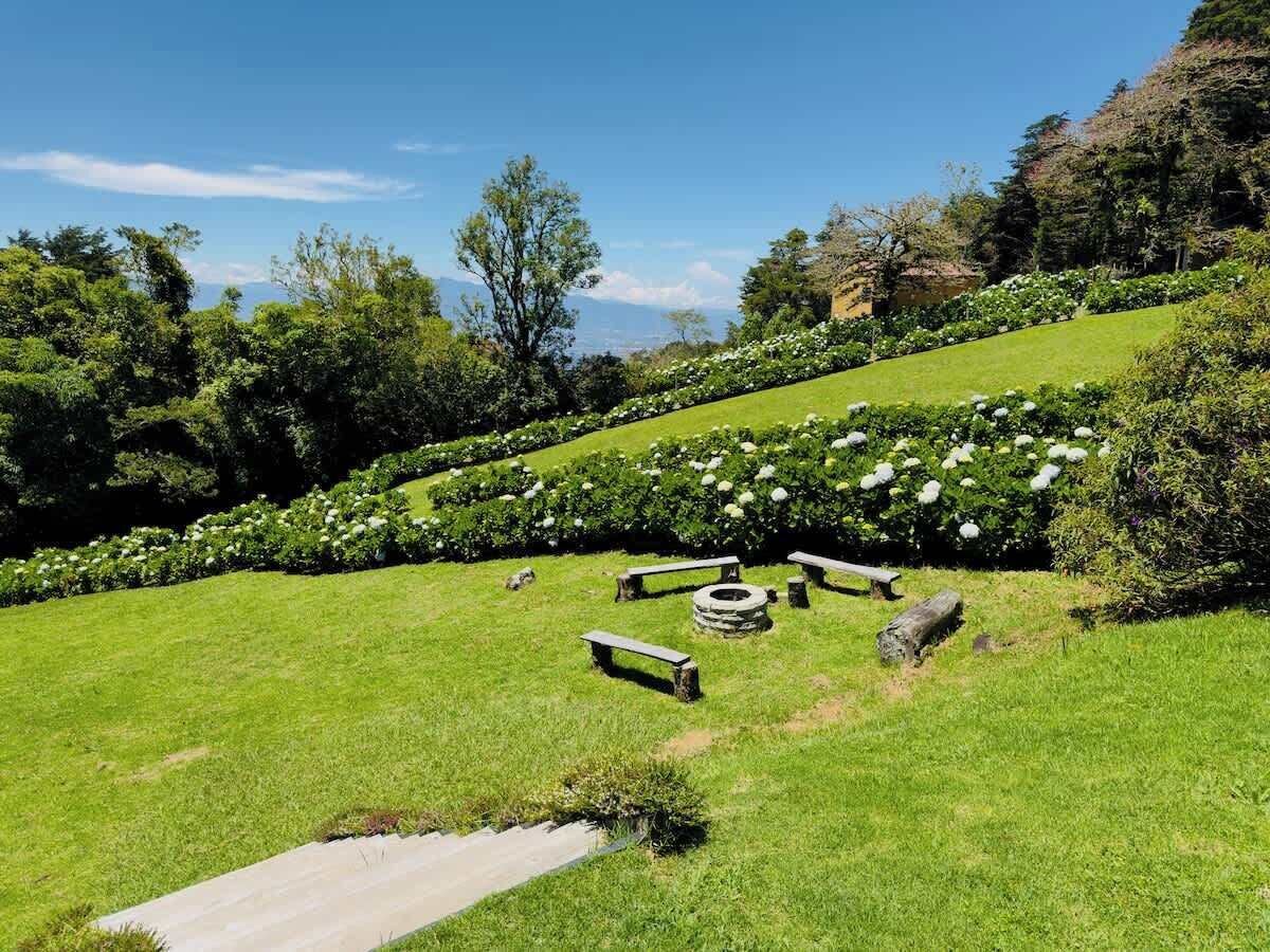 A well-maintained grassy area with stone benches and a circular stone structure, surrounded by lush greenery and trees.