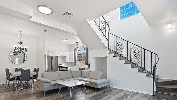 A modern living room with a sectional sofa, dining table, and chairs. A staircase with a decorative railing leads to an upper level.
