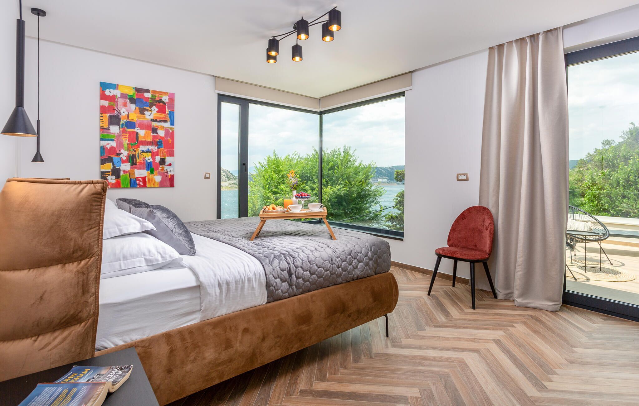 Ein modernes Schlafzimmer mit einem großen Bett, einem Esstisch mit Frühstück, einem roten Sessel und Blick ins Freie.
