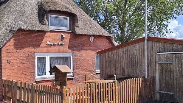 Une maison traditionnelle à toit de chaume, dotée d’une façade en briques, d’une clôture en bois et d’une petite table et d’un banc en bois à l’extérieur.
