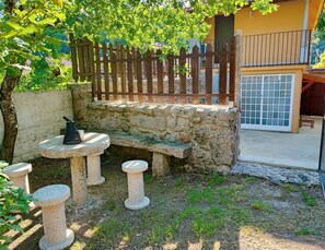 Un banco de piedra con una mesa y taburetes en un jardín, rodeado por una valla de madera y un muro de piedra.