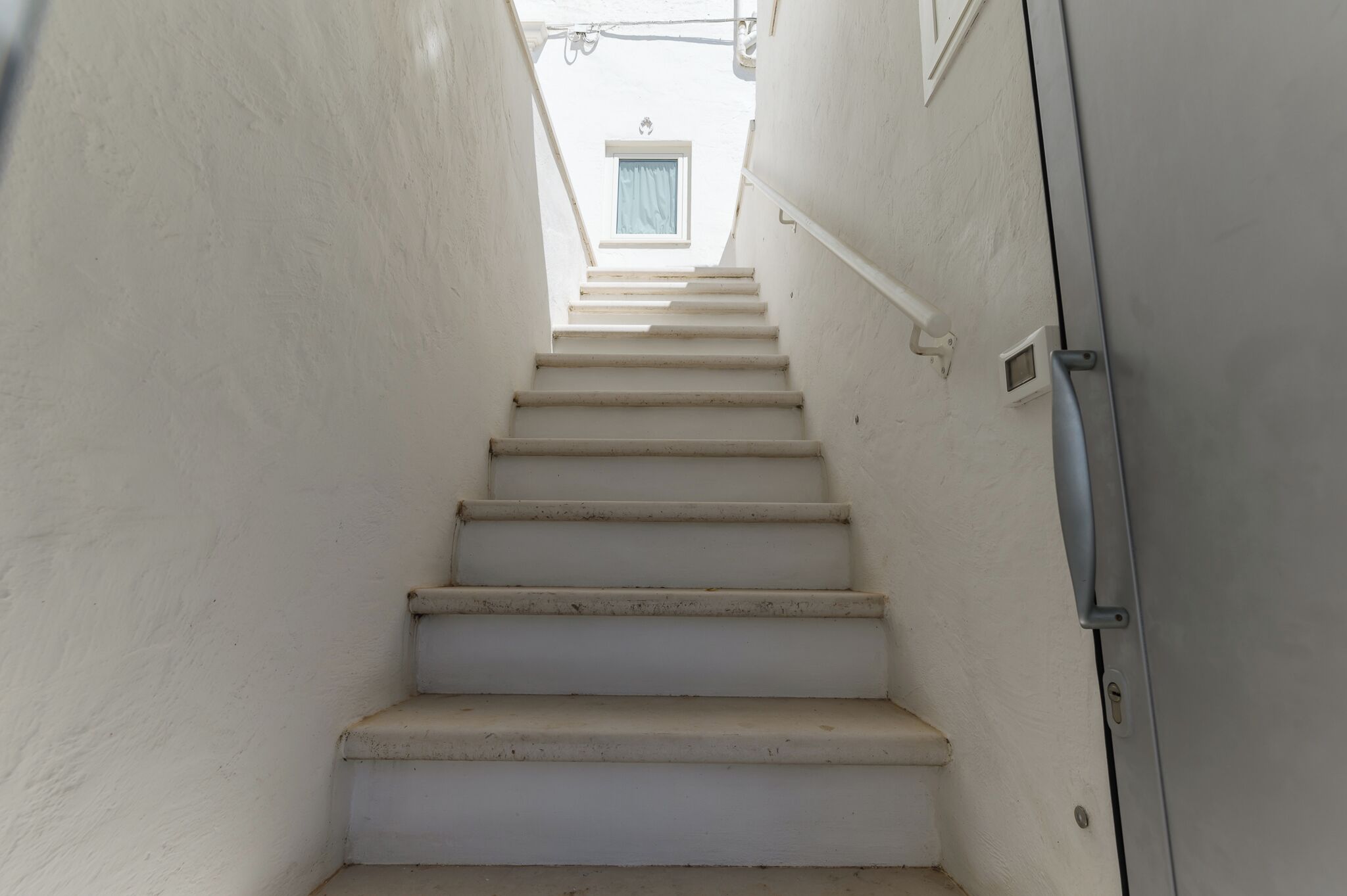 Un escalier étroit aux murs blancs mène à une fenêtre ornée d’un rideau bleu.