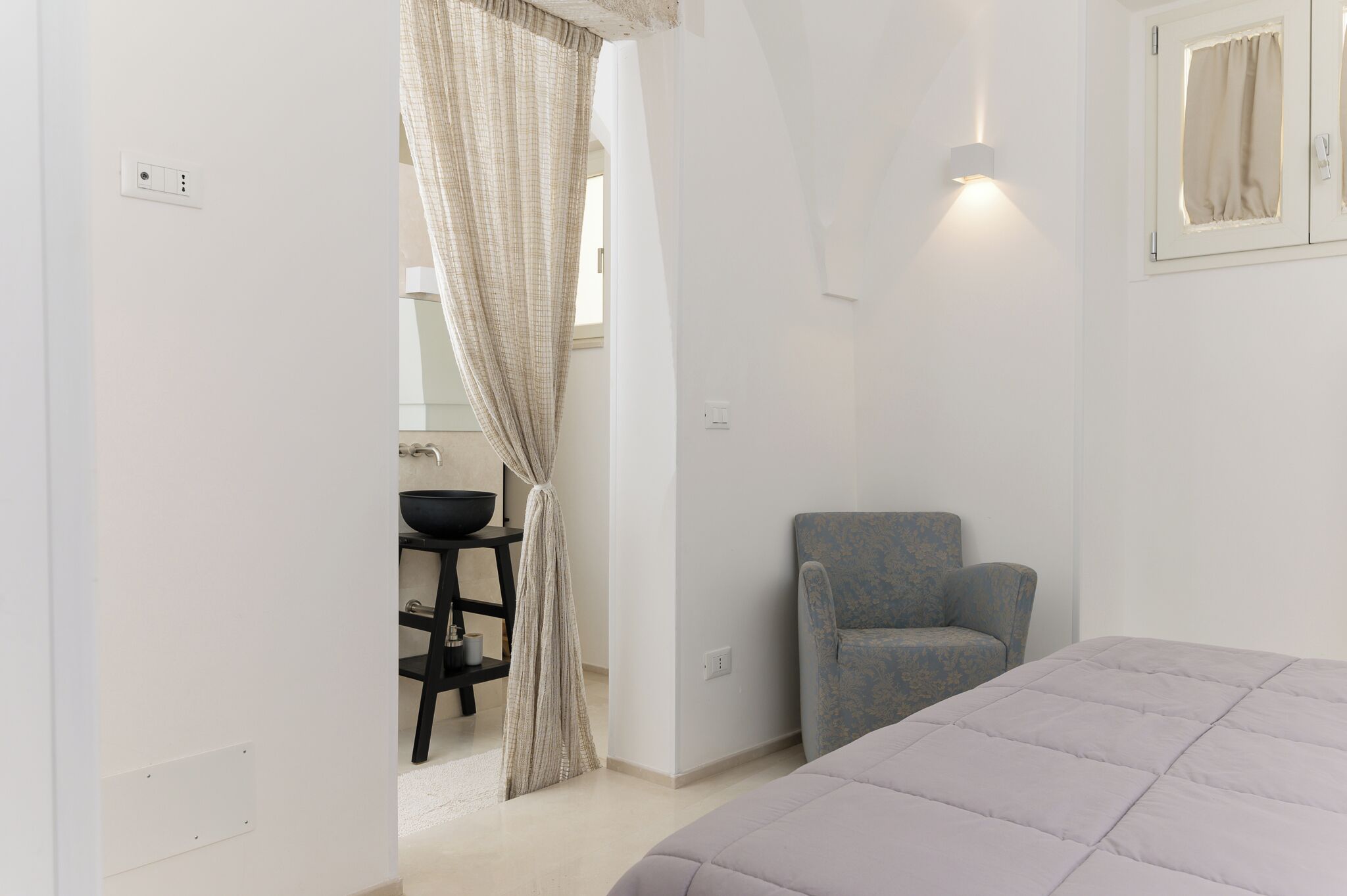 Un dormitorio con cama, una silla, un lavamanos y una lámpara fijada en la pared.