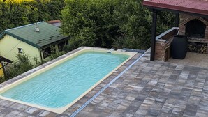 A rectangular swimming pool with a tiled deck, a brick barbecue area, and a covered seating area.