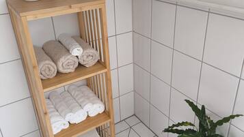 Ein Eckbereich eines Badezimmers mit einem hölzernen Regal, das gerollte Handtücher, eine Topfpflanze und eine weiße Vase beherbergt.
