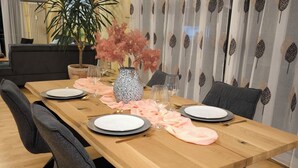 A dining area with a wooden table set for four, surrounded by chairs. A potted plant and a vase with pink flowers are on the table.