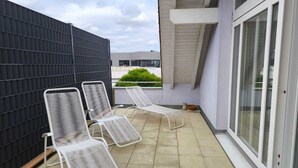 A balcony with three white metal lounge chairs, a black fence, and a view of a building in the background.