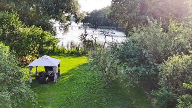 Un belvédère avec une table et des chaises, situé dans un espace verdoyant près d’une étendue d’eau, et entouré d’arbres.