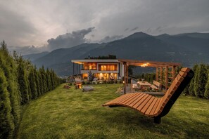 A modern house with a wooden deck and a pergola, surrounded by a well-maintained lawn and tall trees, with mountains in the background. - Ortsried-Hof, Apartment Panorama - Dachterrasse mit Whirlpool und Sauna (Tarsch)