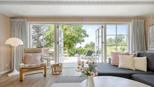 A spacious living room with a large sliding glass door leading to an outdoor area, a comfortable sofa, a chair, and a small table with a vase of flowers. - Amazing home in Ebeltoft with WiFi (Ebeltoft)