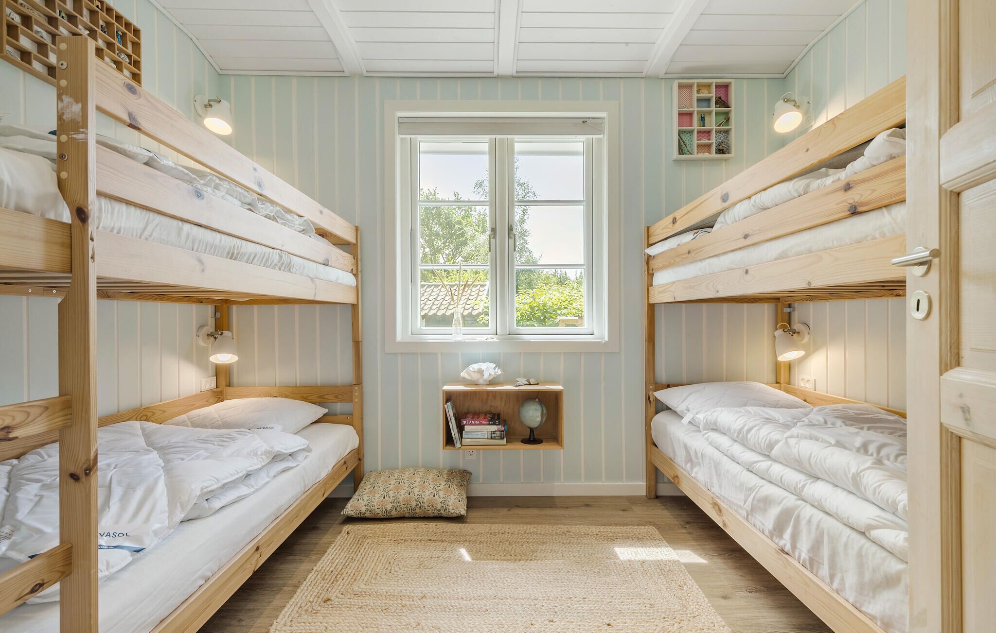 Ein Zimmer mit drei Etagenbetten, einem kleinen Regal mit Büchern, einem Fenster mit Blick auf Grünflächen und einem Teppich auf dem Boden.