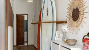 Ein Flur mit einem Surfbrett, das an der Wand lehnt, einem Feuerlöscher und einem dekorativen Spiegel in Sonnenstrahlform.
