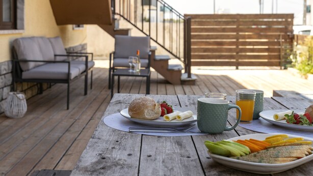 Eine Holzterrasse mit Gartenmöbeln, einem Frühstückstisch mit Essen, einer Tasse und einem Glas Orangensaft.