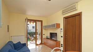 A living room with a sofa, a wooden dining table, and a TV on a cabinet. There is a door and an air conditioning unit on the wall. - Borgo delle Colline 19 by Wonderful Italy (Manerba del Garda)