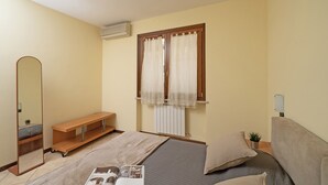 A bedroom with a bed, a bedside table, a mirror, a window with curtains, a radiator, and a shelf. - Borgo delle Colline 19 by Wonderful Italy (Manerba del Garda)