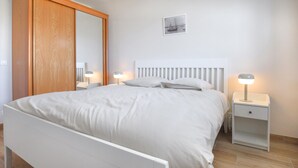 A neatly arranged bedroom with a white bed, two bedside tables with lamps, a wooden wardrobe, and a framed picture on the wall. - 3 bedroom awesome home in Créances (Créances)