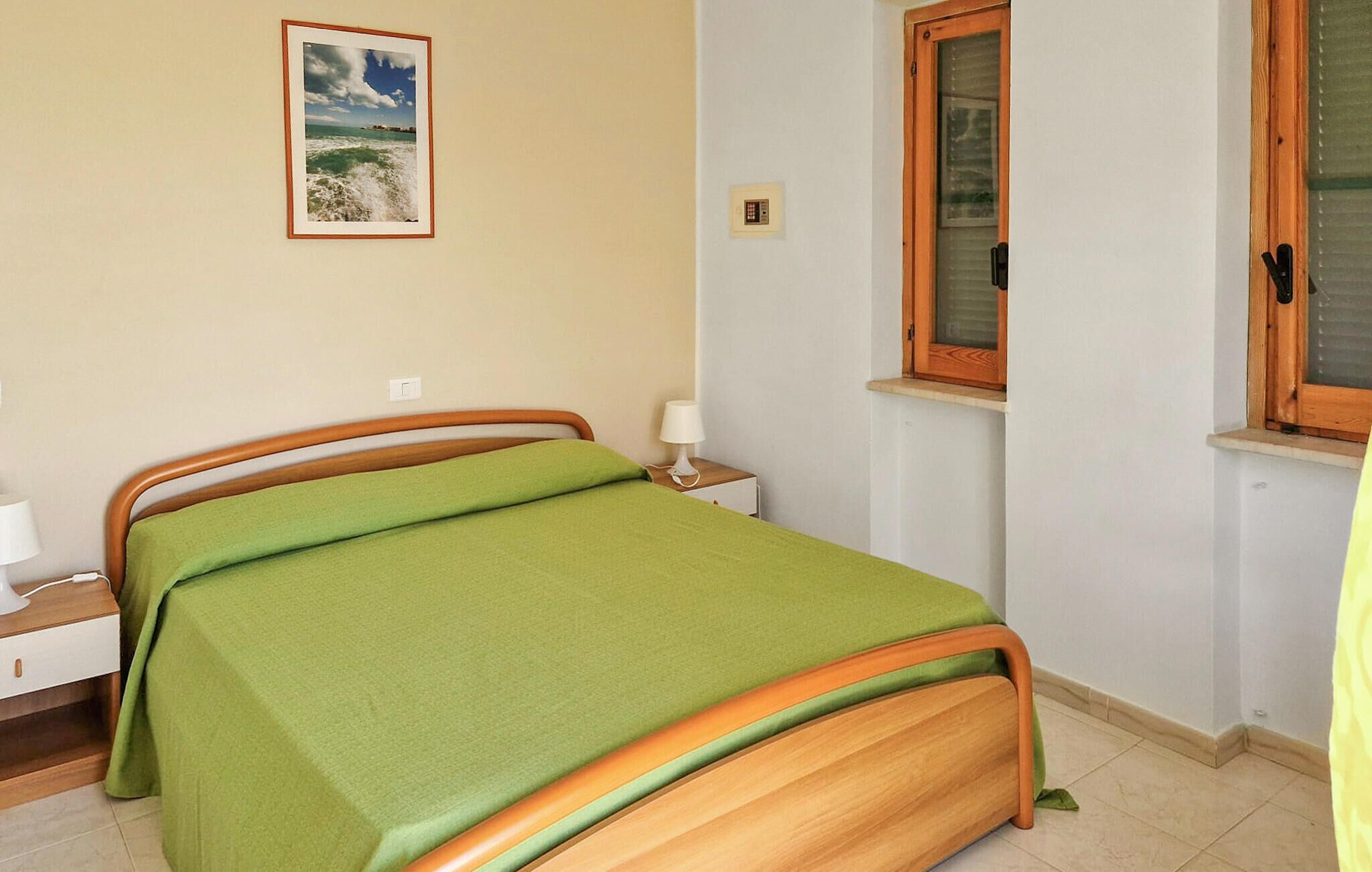 Une chambre à coucher avec un cadre de lit en bois, une literie verte, une table de chevet avec une lampe, une fenêtre avec des stores et un interrupteur fixé au mur.