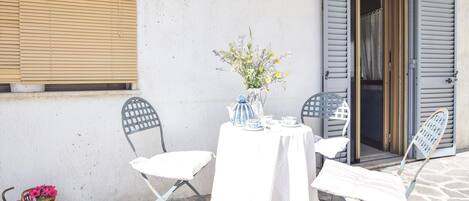 Un tavolo apparecchiato con tovaglia bianca, teiera e tazzine azzurro e bianco, e un vaso con fiori. Due sedie con struttura in metallo blu e cuscini bianchi. Sul pavimento, un cesto di vimini con fiori e una bicicletta.