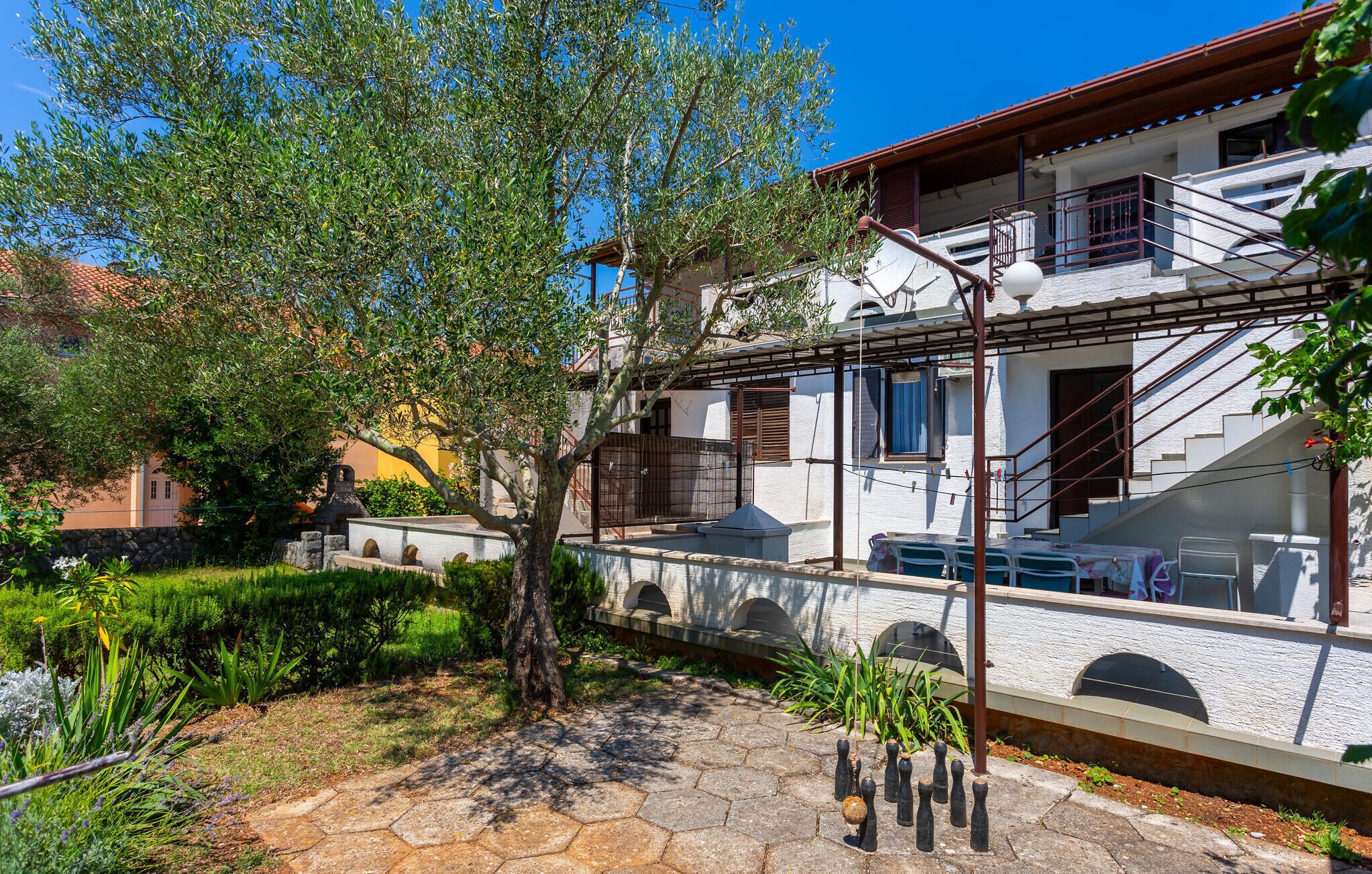 A courtyard with a tree, a stone pathway, and a building with a balcony and stairs.