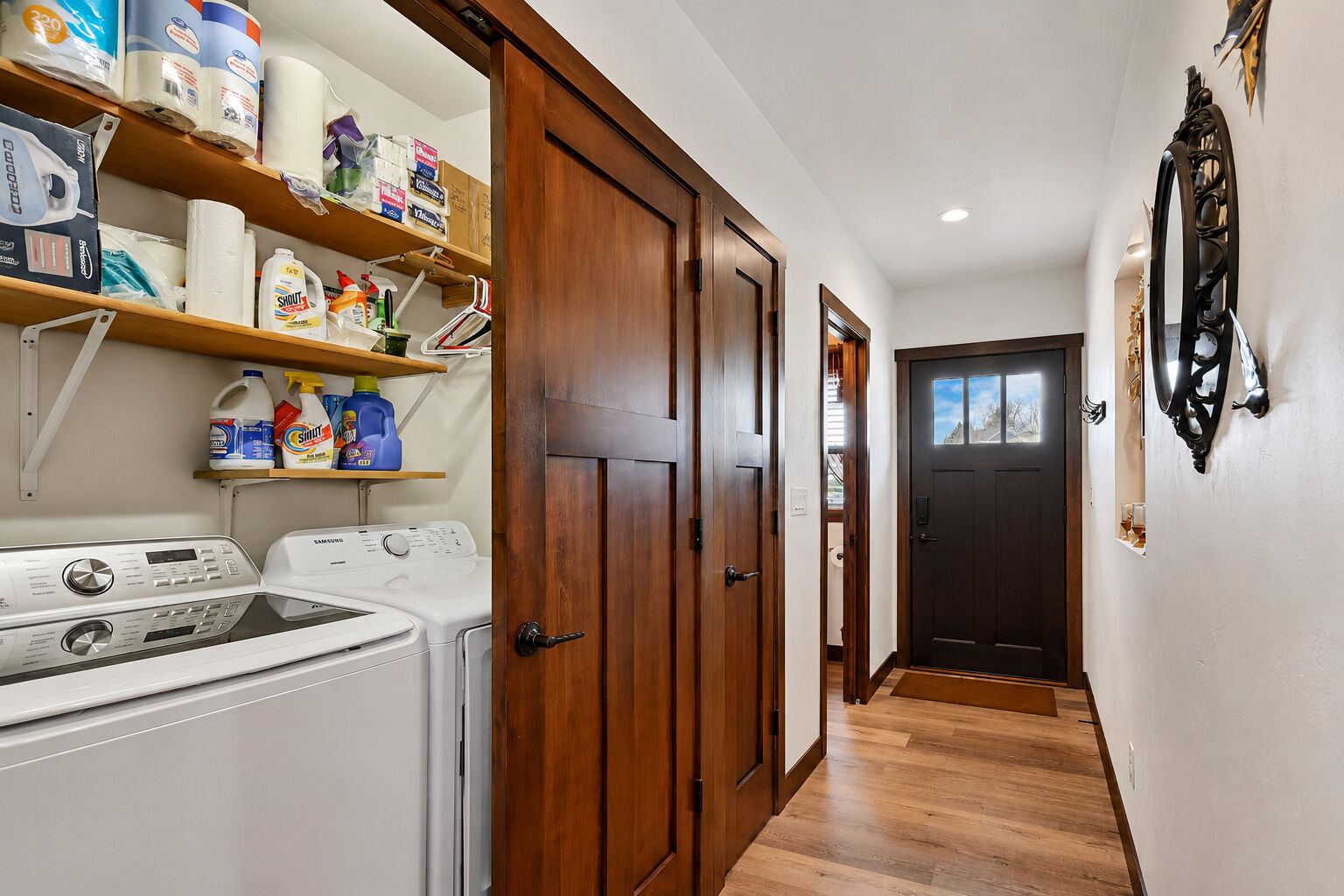 A laundry room with washing machines, shelves with cleaning supplies, and a wooden door leading to another room.
