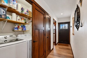 Un lavadero con lavadoras, estantes con productos de limpieza y una puerta de madera que da a otra habitación.