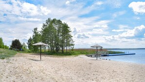 Ein sandiger Strand mit zwei hölzernen Pavillons, einer anlegenden Mole in ruhigem Wasser und einer Baumreihe im Hintergrund.
