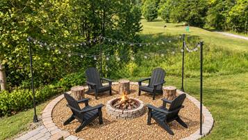 A circular outdoor seating area with Adirondack chairs and a fire pit, surrounded by a brick border and string lights.