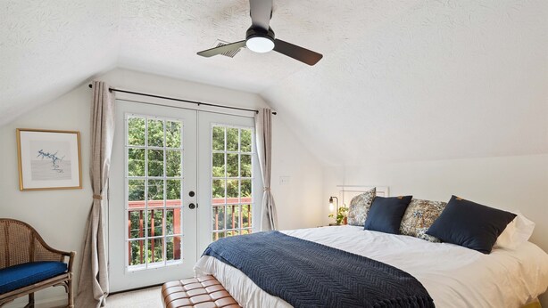 Un dormitorio con cama, un banco, una silla, un ventilador de techo y una ventana con cortinas.
