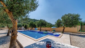 Ein rechteckiger Swimmingpool mit gefliestem Sonnendeck, umgeben von Olivenbäumen und blauem Himmel.