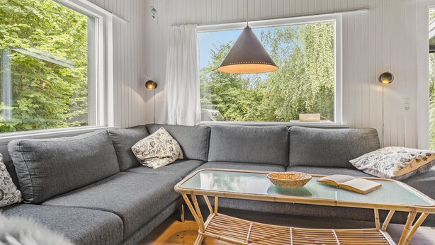 Ein modernes Wohnzimmer mit einem grauen Ecksofa, einem Glastisch und einem großen Fenster mit Blick auf Grünflächen.