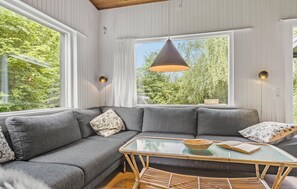 Un salon moderne avec un canapé d’angle gris, une table basse en verre et une grande fenêtre donnant sur un paysage verdoyant.