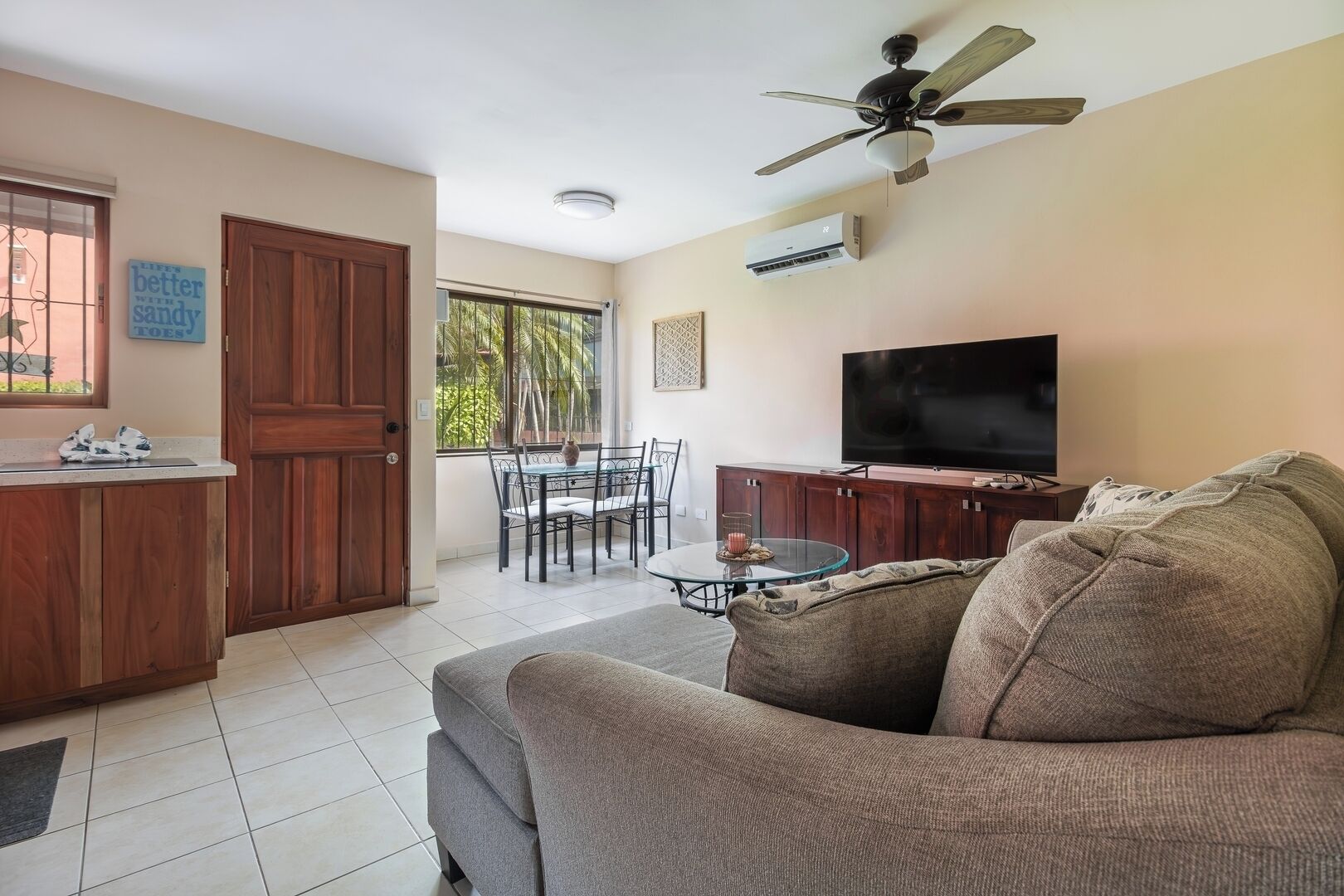 A living room with a sofa, TV, ceiling fan, dining table, chairs, and a kitchen area with a wooden cabinet and an air conditioner.