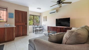 A living room with a sofa, TV, ceiling fan, dining table, chairs, and a kitchen area with a wooden cabinet and an air conditioner.