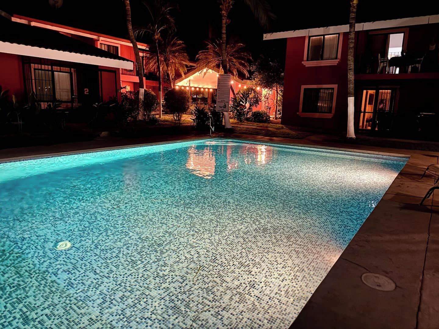 Una piscina con superficie de azulejos, rodeada de edificios con balcones y palmeras.