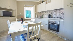 Une cuisine moderne avec des meubles blancs, une table à manger avec des chaises, un four encastré et une fenêtre avec des volets bleus.