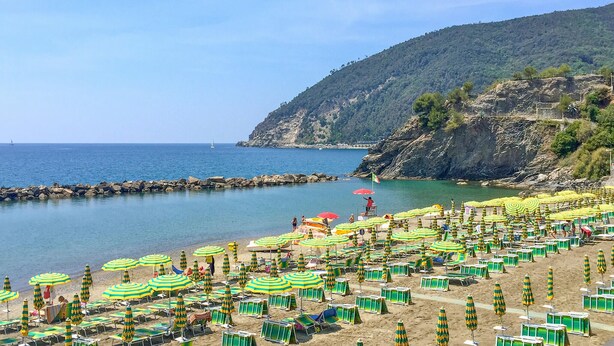 Une plage parsemée de nombreux parasols et de chaises longues, un littoral rocheux et un voilier à l’horizon.