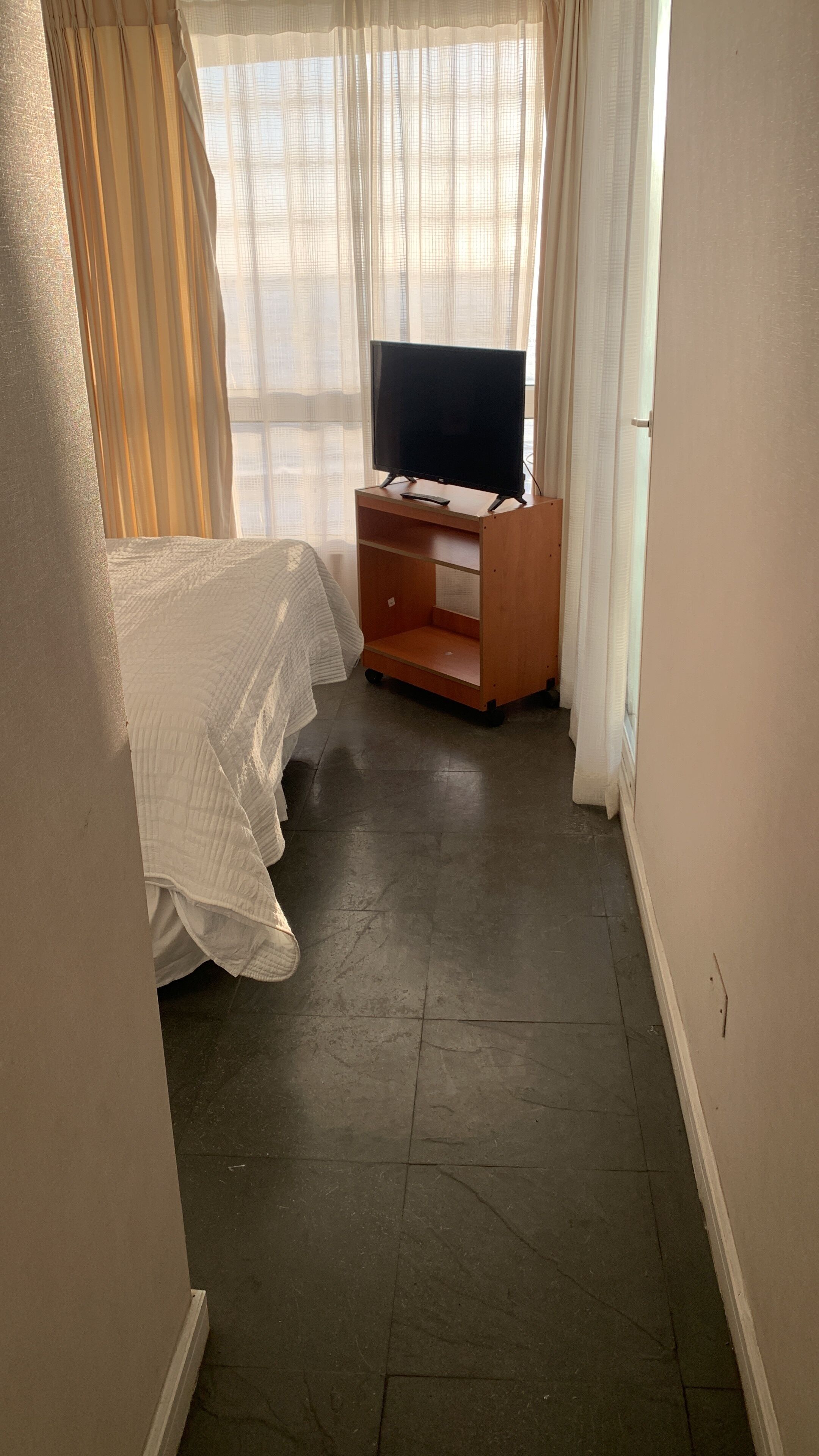 Une pièce comprenant un lit, un meuble en bois pour la télévision et une fenêtre orn ée de rideaux légers.