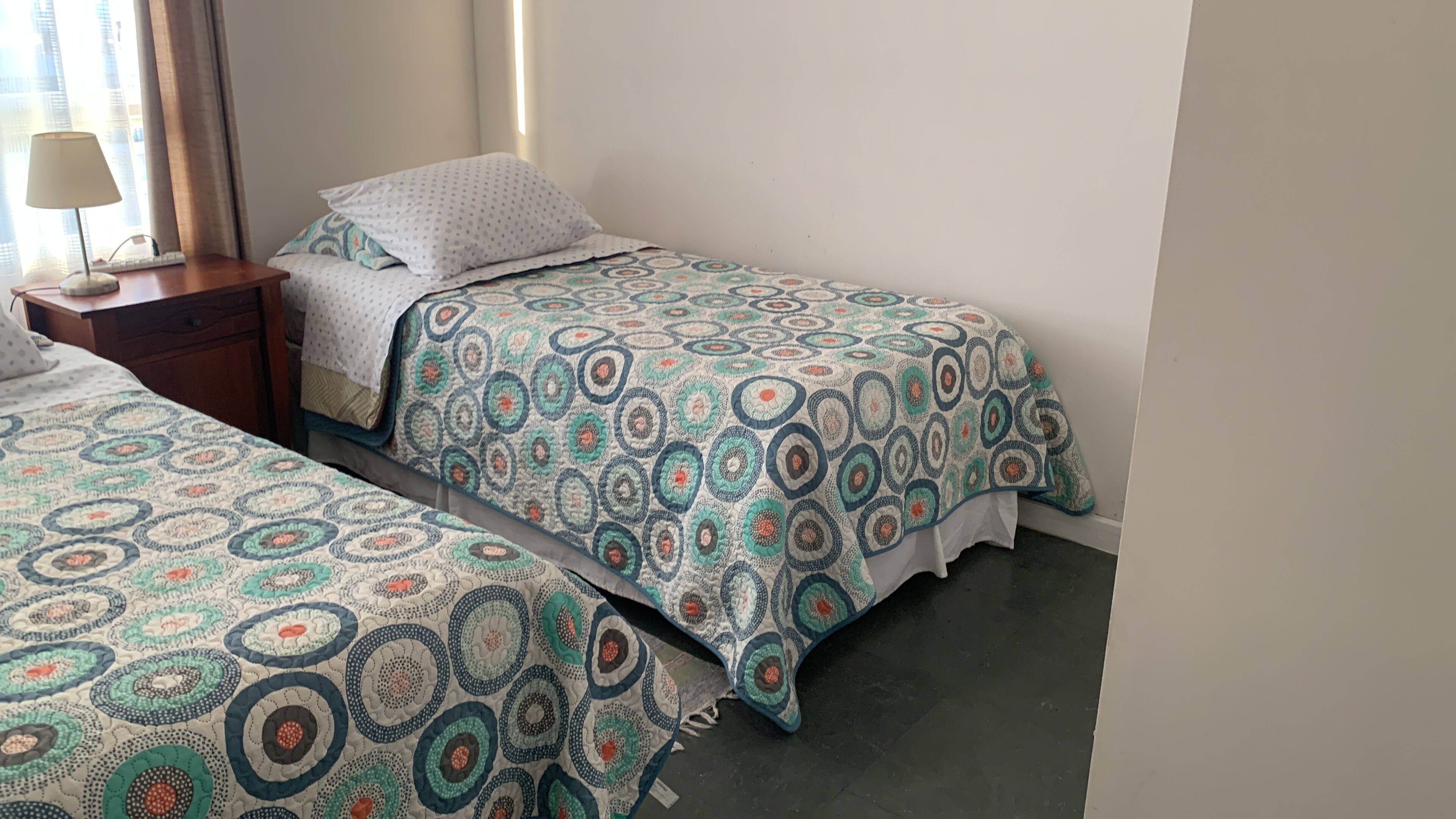 Een hotelkamer met twee eenpersoonsbedden, elk met een beddekleed met een patroon en een kussen. Er staat een nachtkastje met een lamp en een klein lade.