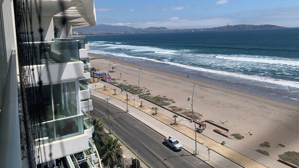 A coastal view from a high-rise building overlooking a beach with a road and a distant city skyline. - La Serena Avenida Del Mar 3 Bedrooms beach view  (La Serena)