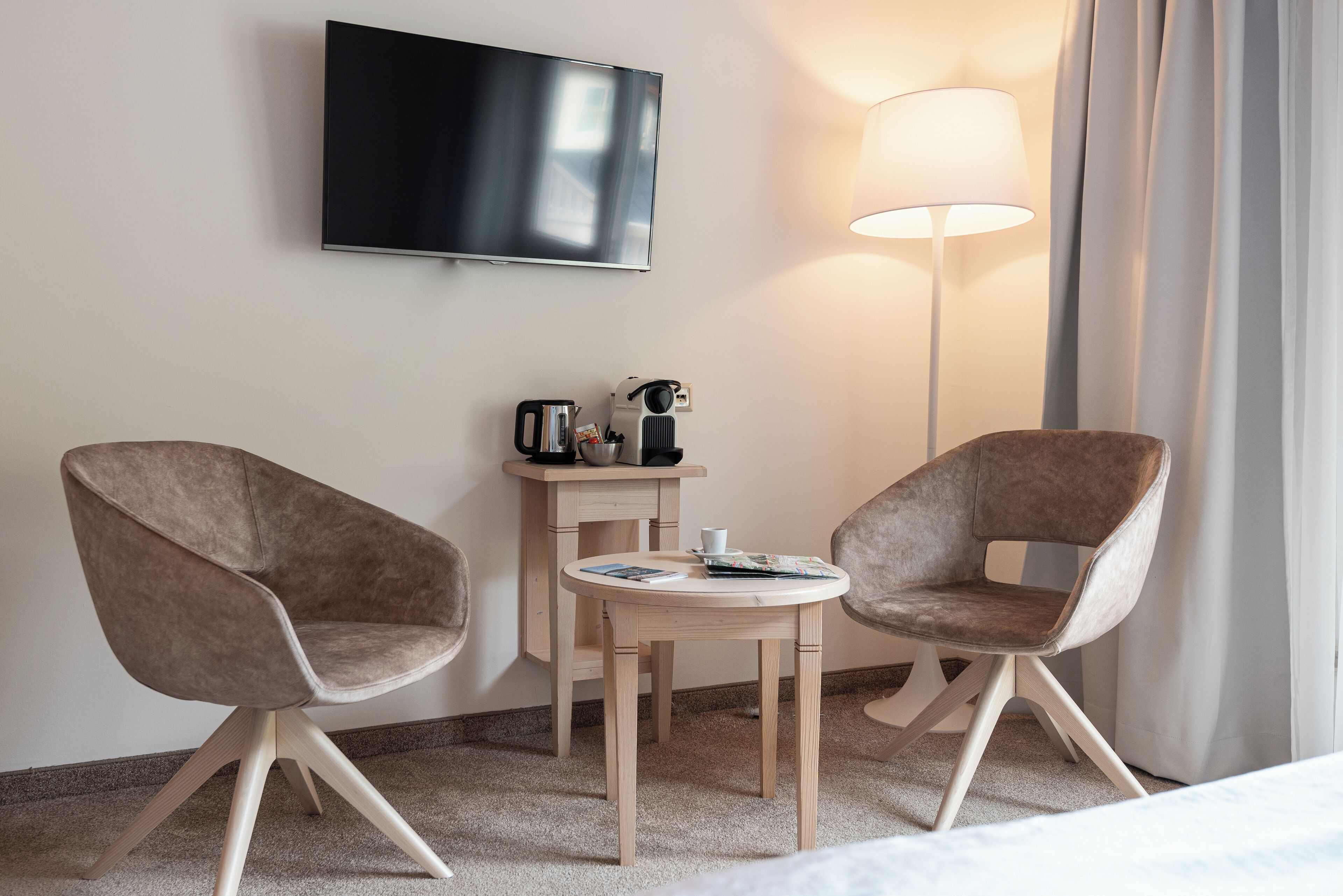 A hotel room with two armchairs, a small wooden table, a flat-screen TV mounted on the wall, a floor lamp, and a coffee machine.