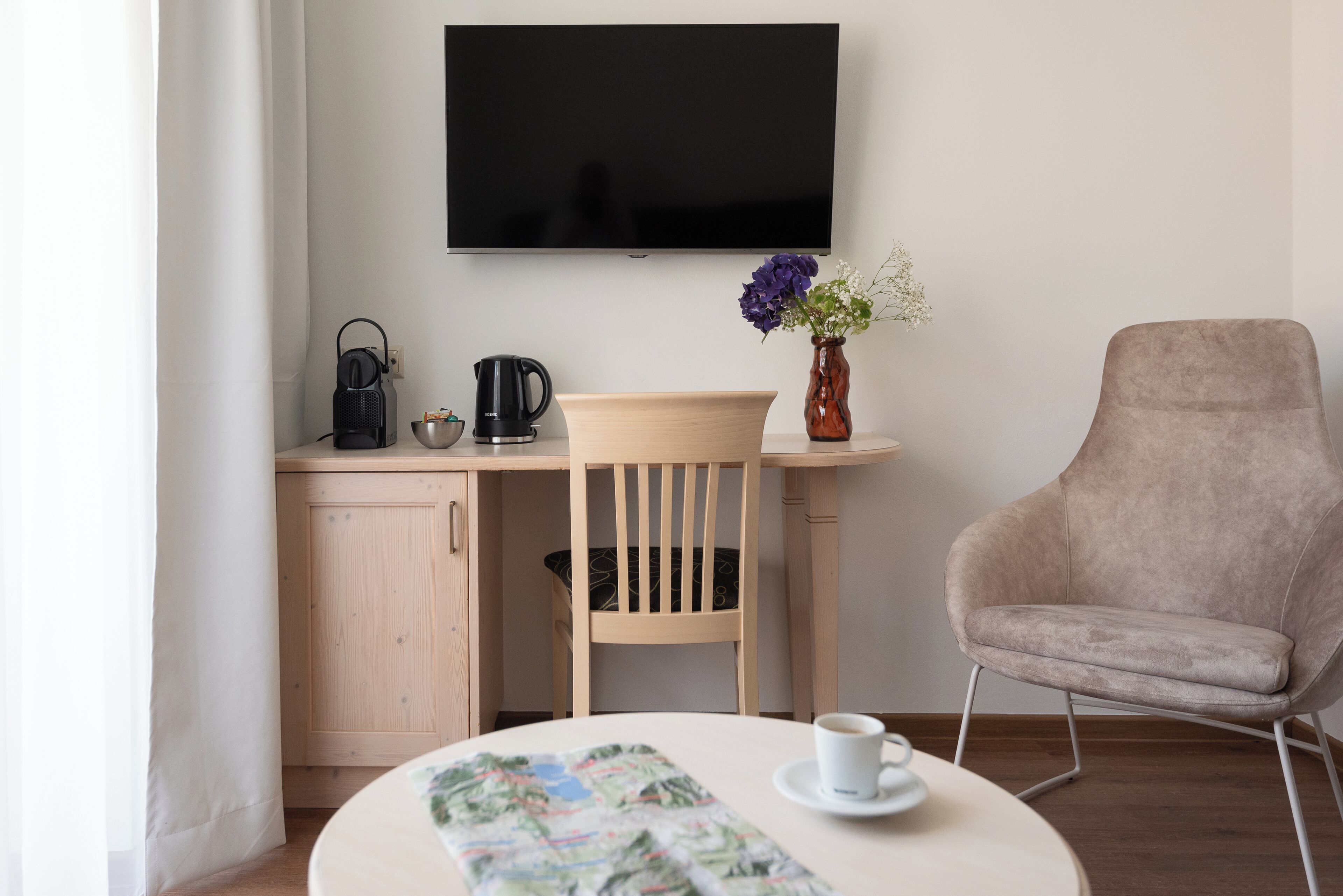 Una habitación moderna con una mesa redonda, una silla, un televisor, una cafetera y un florero con flores.