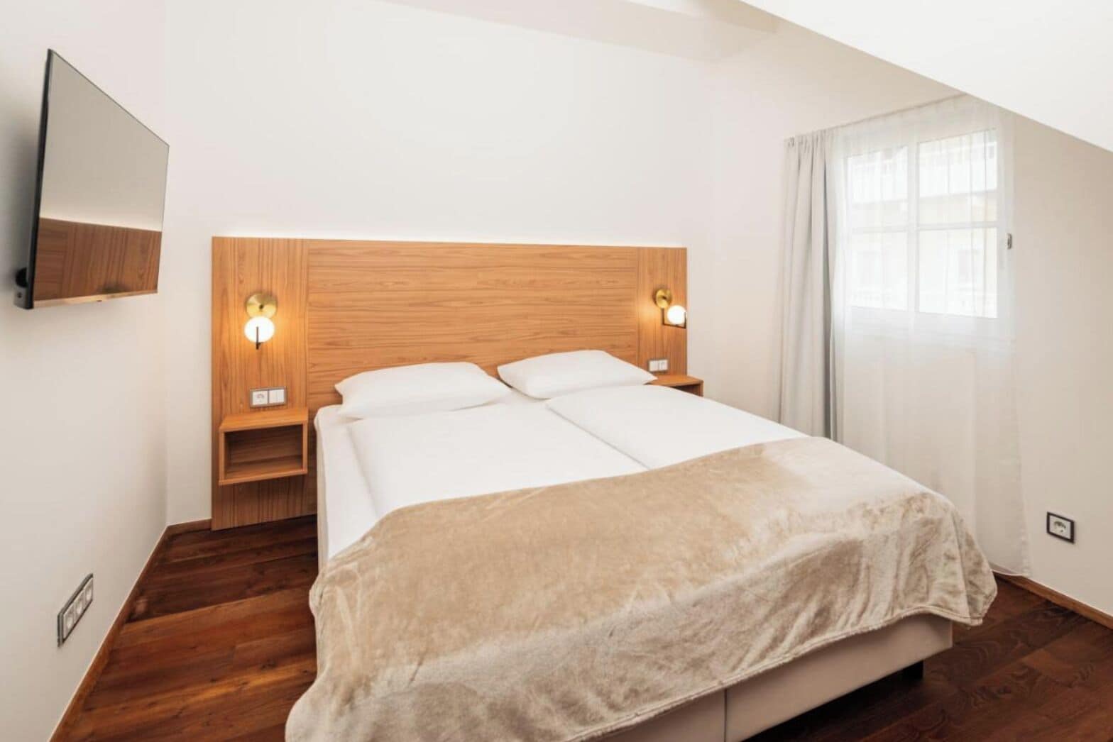 A modern hotel room with a wooden headboard, two bedside tables with lamps, a large bed with a beige blanket, and a large window with curtains.