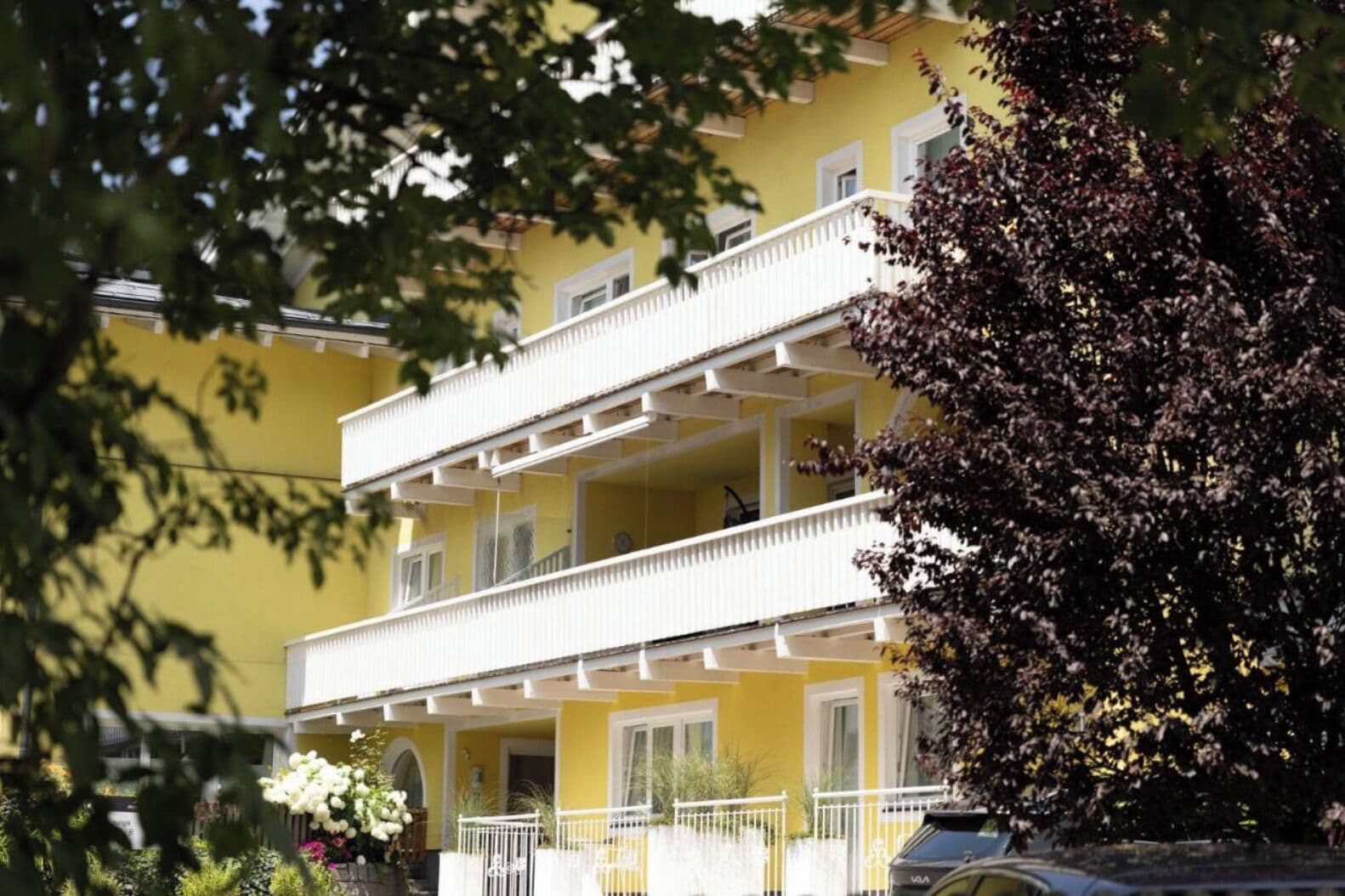 Un immeuble jaune à deux étages, doté d’un balcon blanc, entouré de verdure et avec une voiture garée à proximité.