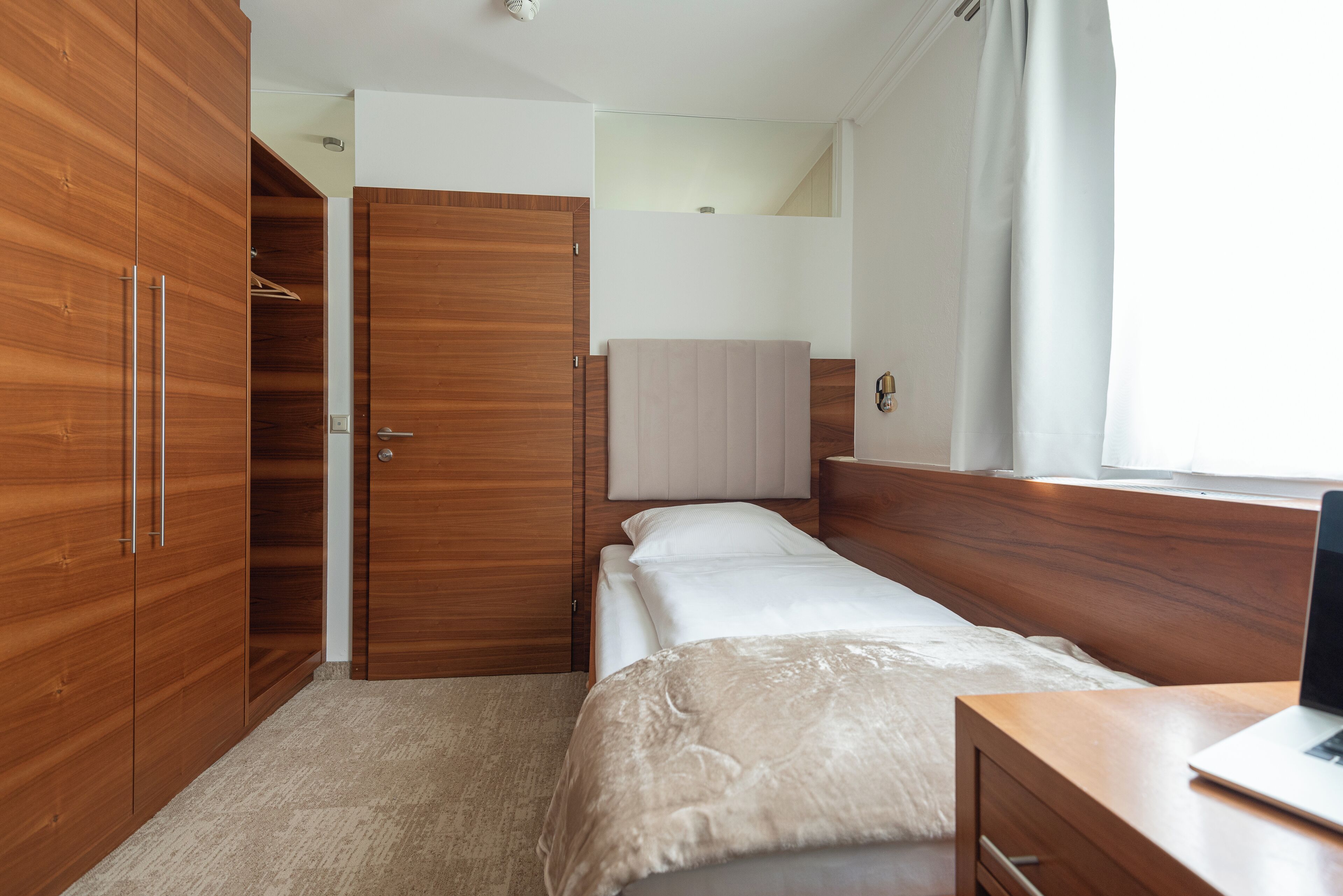 A bedroom with a bed, wooden wardrobe, and a desk with a laptop.