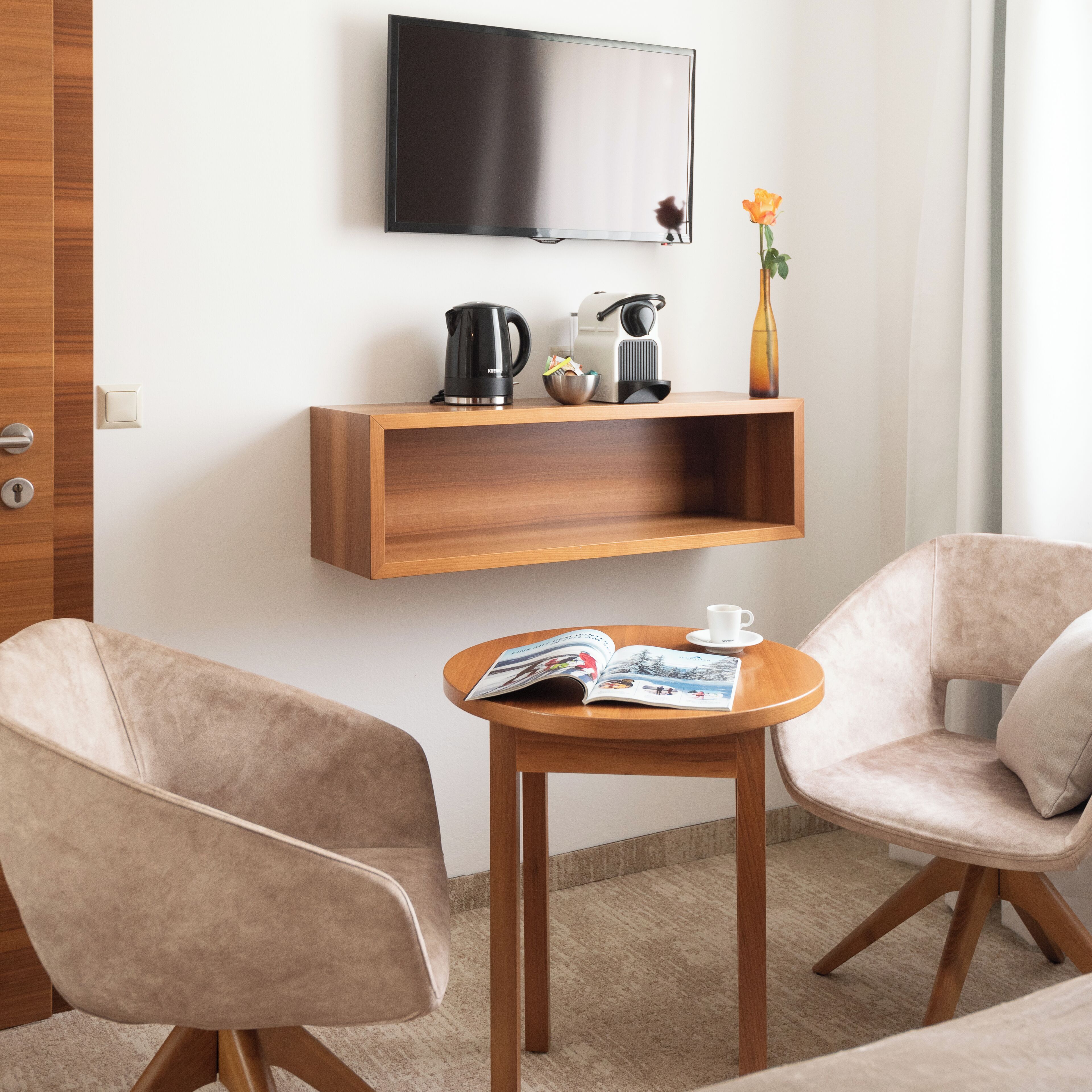 A modern hotel room with a wooden TV stand, a coffee maker, a kettle, and a vase with a single flower. There are two beige armchairs and a small wooden table with a magazine.
