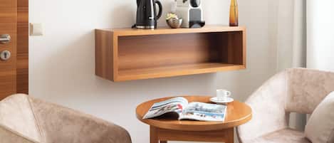 A modern hotel room with a wooden TV stand, a coffee maker, a kettle, and a vase with a single flower. There are two beige armchairs and a small wooden table with a magazine.