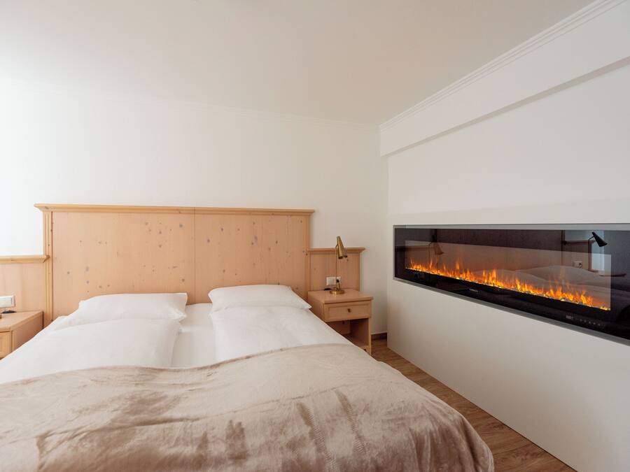 Quarto com cama, mesas de cabeceira, um aparador, lareira na parede e televisão.