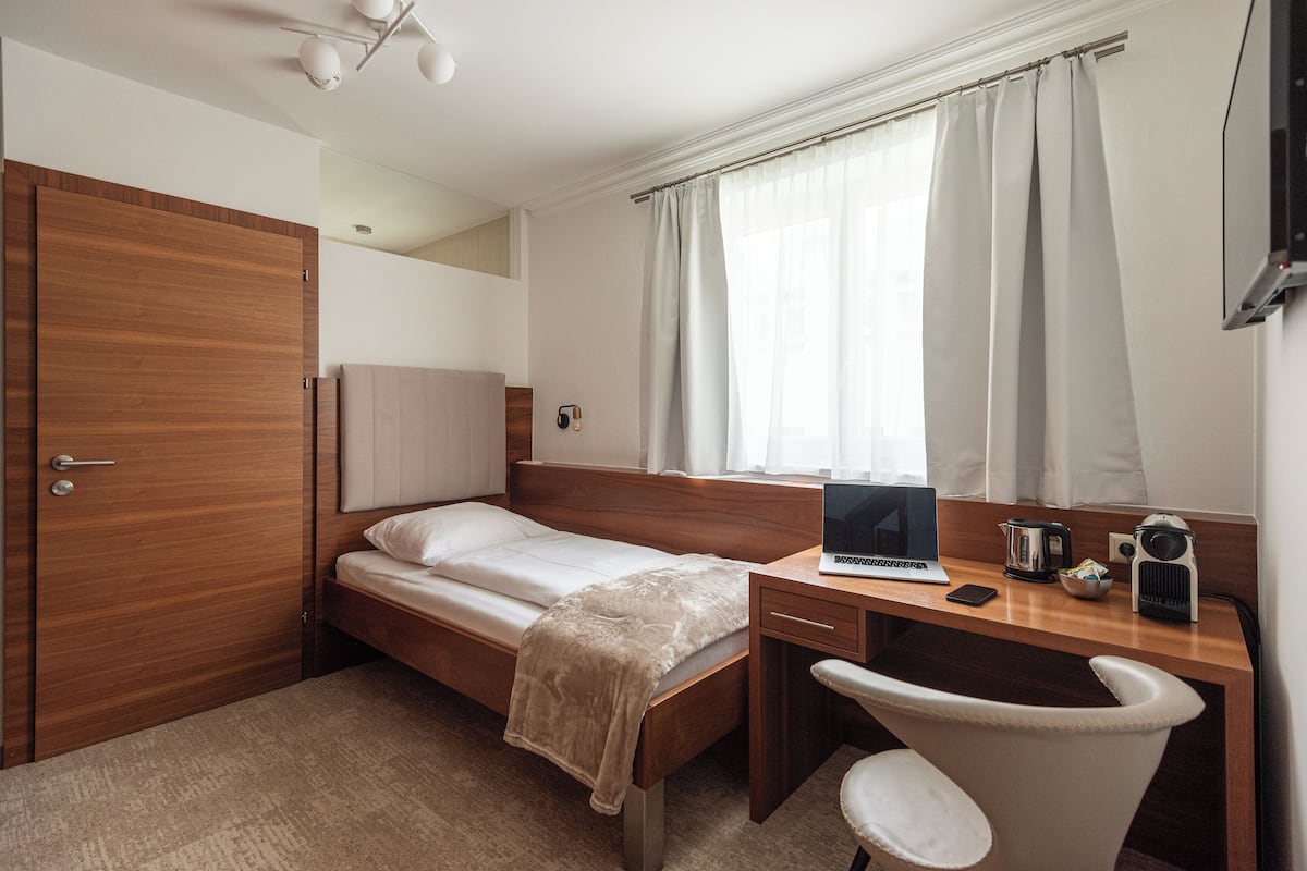 A bedroom with a wooden bed, a desk with a laptop, a chair, and a window with curtains.