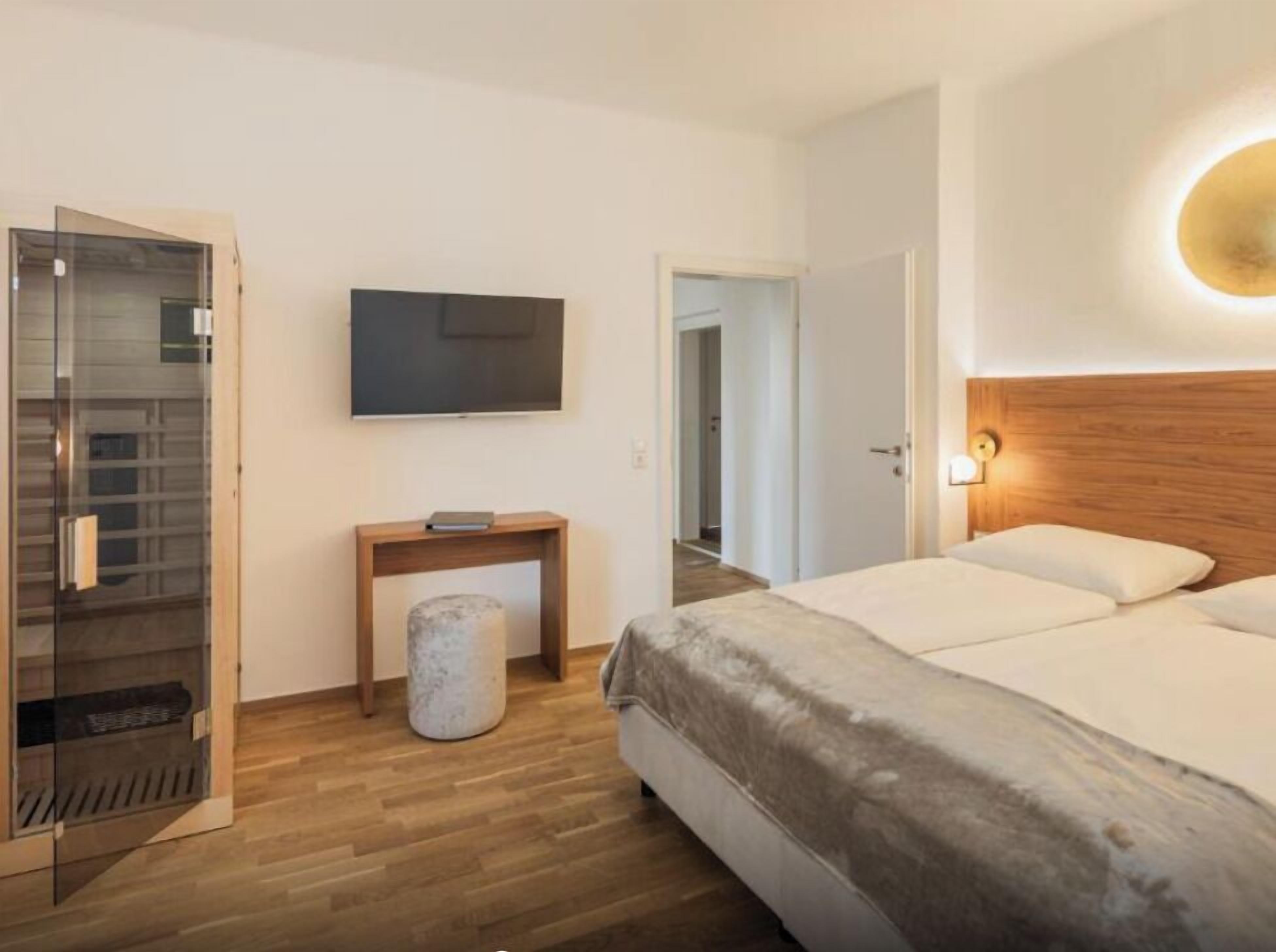 A hotel room with a bed, a TV mounted on the wall, a wooden headboard, and a small bedside table with a lamp.